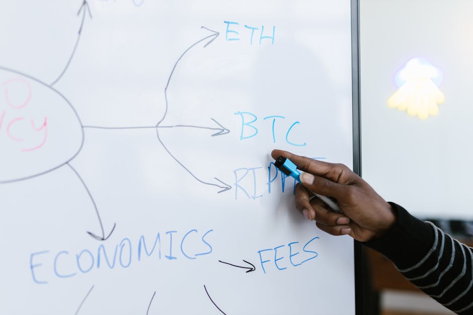 A person's hand holding a blue marker is seen writing on a whiteboard, where various cryptocurrency abbreviations such as ETH, BTC, and XRP are written in blue, with arrows pointing to the words 'FEES' and 'ECONOMICS'. The whiteboard includes a diagram or flowchart related to cryptocurrency economics and transaction costs, with a metal frame and a partially visible decorative light or sticker on the right side. The setting appears to be an indoor environment, possibly an office or classroom focused on digital currency discussions. The hand is positioned near the middle of the board, emphasizing the process of explaining or illustrating cryptocurrency transaction fees and economic concepts, which subtly aligns with content about moving or logistics costs in a professional context, reflecting careful planning and process management in house removals by Lambeth Removals.