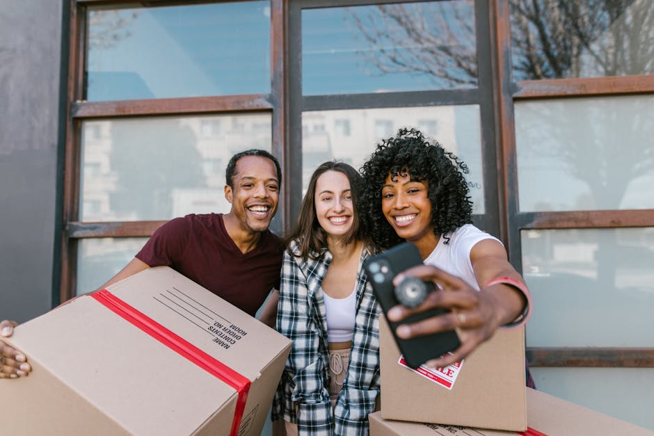 Three individuals standing outdoors in front of a building with large windows, smiling as they pose for a selfie. The person on the right, a woman with curly black hair wearing a white t-shirt, holds a smartphone and a cardboard box with a red packing label. The man on the left, with short dark hair and wearing a dark red t-shirt, is holding a cardboard box wrapped with red tape, labeled 'Home Contents'. The woman in the middle, with long brown hair and a plaid shirt, also holds a cardboard box with packing labels. They appear to be preparing for home relocation, with the background suggesting an urban environment. The scene reflects packing and moving activities, with bags and moving supplies around, indicating a furniture transport or moving process. It is a bright day, and the group is engaging in a friendly, celebratory moment related to their move, which could be assisted by [COMPANY_NAME]’s removals services.