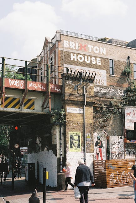 A city street scene showing a brick building with multiple layers of exterior walls, featuring large painted signs including the words 'BRIXTON HOUSE' and various graffiti and posters. A metal fire escape with stairs extends from the building's upper floors, alongside some plant growth along the facade. Below, pedestrians walk along the sidewalk, some carrying bags, with a distinct yellow and black striped barrier on the left supporting an elevated roadway or railway. The street environment is urban, with a mix of commercial and residential elements, and the lighting suggests daytime with partly cloudy skies. The scene relates to house removals and relocating services provided by Lambeth Removals, illustrating aspects of moving logistics like loading or transporting furniture and belongings within a typical Brixton neighbourhood.