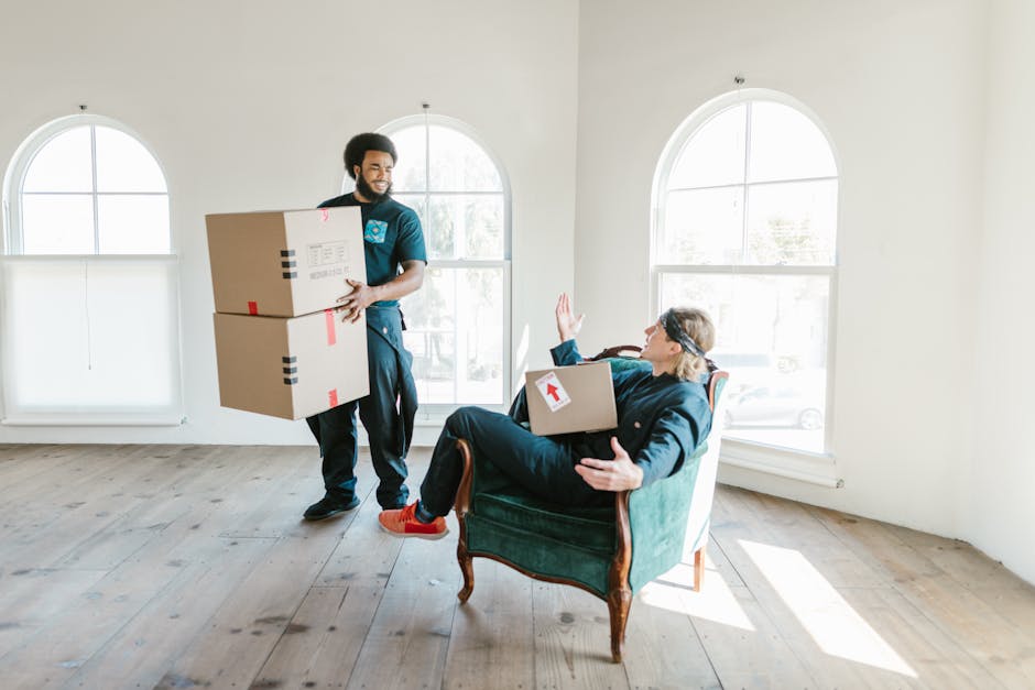 A professional removal team member from Lambeth Removals is inside a bright, spacious room with large arched windows allowing natural light to illuminate the space. The team member, dressed in a uniform, is carrying two cardboard boxes stacked on top of each other, with packing tape securing the lids. Inside the room, a woman is seated on a vintage-style armchair with wooden legs and green upholstery, holding a cardboard box with a red arrow label on it, while raising her right hand in a greeting or signal gesture. The room has a light wooden floor, and the walls are painted white, creating a clean and minimal environment suitable for packing and home relocation activities. The scene captures the process of furniture transport and packing during a house move, with the removal service set up for loading or unloading items in preparation for transport. Lambeth Removals’ involvement in home removals and moving logistics is subtly implied through the professional handling of boxes and equipment visible in the space.
