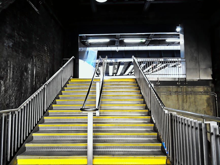 Interior view of a staircase inside a building used for house removals, featuring metal handrails on both sides and yellow safety strips along the edge of each step. The staircase is illuminated by overhead lighting, with a dark, textured wall on the left and a metallic door and railing at the top. The steps appear to be made of metal or a similar durable material suitable for furniture transport and moving operations. This environment suggests a stair access point for a flat or apartment, relevant for planning home relocation logistics with Lambeth Removals, as shown on the webpage about small flat removals and stair access advice.