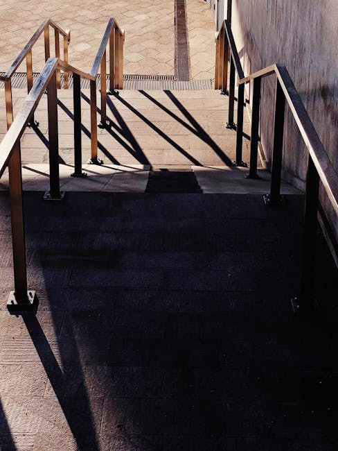 The image shows a staircase inside a residential property, with a marble or stone ramp providing stair access. The staircase features a dark wooden handrail and white balustrades, leading up to a small landing with several potted plants of various sizes and types placed on a windowsill. The window allows natural light to illuminate the area, highlighting the potted greenery and the textured surface of the stairs. To the left of the stairs, a tall, silver ladder leans against the wall, likely used during packing or furniture protection activities during a house move. The floor at the base of the stairs has a decorative patterned tile design in muted tones. A soft, warm light fixture hangs from the ceiling, adding ambient illumination. This scene captures a typical home environment where furniture and boxes are prepared for transport, supporting the context of professional removals and relocating furniture, as provided by Lambeth Removals for small flat removals with stair access challenges.