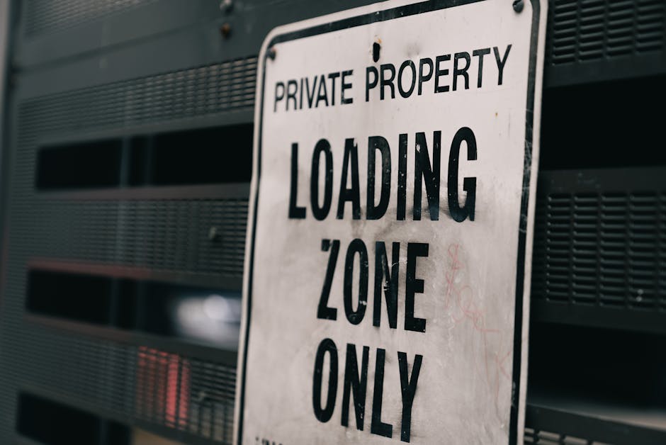 A close-up view of a white metal sign attached to a dark grey or black vented building exterior. The sign displays the text 'PRIVATE PROPERTY LOADING ZONE ONLY' in bold, black capital letters. The background shows horizontal slats or louvers, likely part of a loading dock or building facade, with some areas slightly out of focus to emphasize the sign. The setting suggests a designated area for moving vehicles and furniture transport, relevant to home relocation and packing and moving services, with natural lighting highlighting the sign's details. This visual context aligns with logistical aspects of professional removals, such as loading and unloading furniture and boxes, as handled by Lambeth Removals, supporting services related to removals and vehicle access permissions, consistent with the information on the Lambeth council parking permits page.