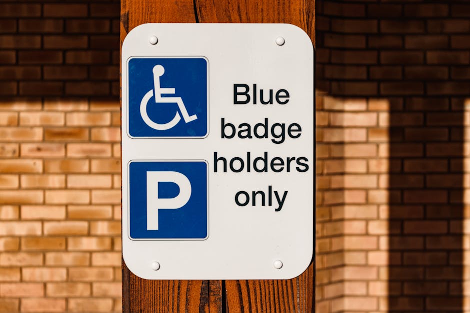 A rectangular white parking sign attached to a wooden post with four screws, displaying a blue handicap symbol and a blue parking symbol. The sign has black text that reads 'Blue badge holders only' next to the respective symbols. The background features a brick wall with horizontal reddish-brown bricks, and sunlight casting shadows on the wall and post. The sign is positioned in an outdoor environment, indicating designated parking areas for individuals with mobility impairments, which is relevant for moving and home relocation services provided by Lambeth Removals.