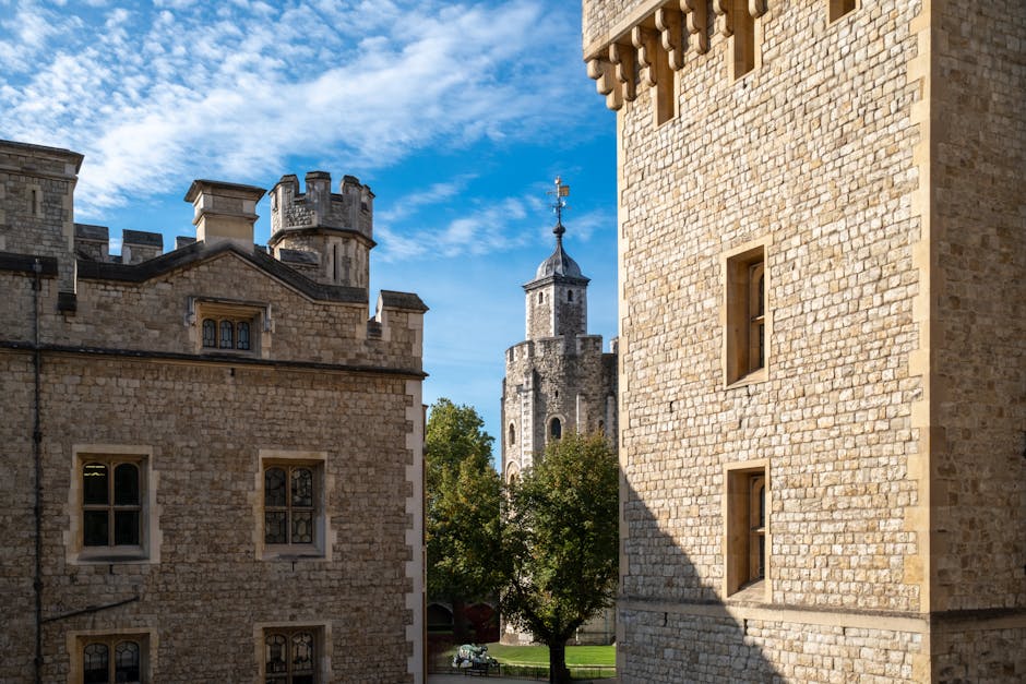 A photograph of historic stone buildings with medieval architectural features, including small rectangular windows and crenellated battlements, captures the exterior of Lambeth Palace near Waterloo Station. In the background, a tall, narrow tower with a conical roof and weather vanes is visible, surrounded by lush green trees. The sky above is partly cloudy with bright blue and white clouds. This scene is taken from a vantage point between two large stone structures, highlighting the textured, beige-colored brickwork of the buildings. The image reflects the historical setting and architectural character, relevant to house and building removals or relocation services offered by Lambeth Removals, as they facilitate the careful transport of properties’ furniture, boxes, and appliances between locations.