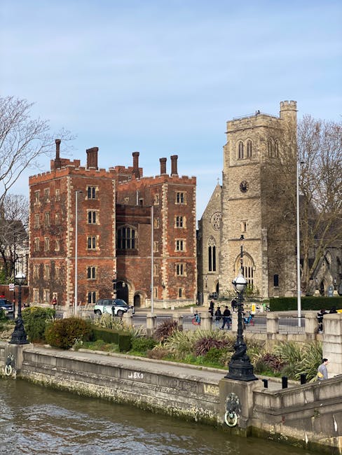 A historic stone and brick building with battlements and multiple chimneys, situated next to a stone church with a tall tower and arched windows, located on a riverside promenade. The promenade features a low stone wall with decorative lamp posts and a paved walkway, with a few pedestrians walking or standing nearby. Vehicles are parked along the street adjacent to the building. The scene is set under a clear sky with some scattered clouds, and leafless trees are visible in the background, indicating a possible early spring or late autumn setting. The environment appears calm and well-maintained, suitable for casual strolling or sightseeing, and is relevant to the context of home relocation and moving services provided by Lambeth Removals, who may handle belongings in such historic residential or commercial properties during packing and moving operations.