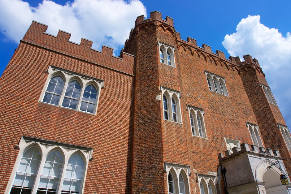 A photograph of historic stone buildings with medieval architectural features, including small rectangular windows and crenellated battlements, captures the exterior of Lambeth Palace near Waterloo Station. In the background, a tall, narrow tower with a conical roof and weather vanes is visible, surrounded by lush green trees. The sky above is partly cloudy with bright blue and white clouds. This scene is taken from a vantage point between two large stone structures, highlighting the textured, beige-colored brickwork of the buildings. The image reflects the historical setting and architectural character, relevant to house and building removals or relocation services offered by Lambeth Removals, as they facilitate the careful transport of properties’ furniture, boxes, and appliances between locations.