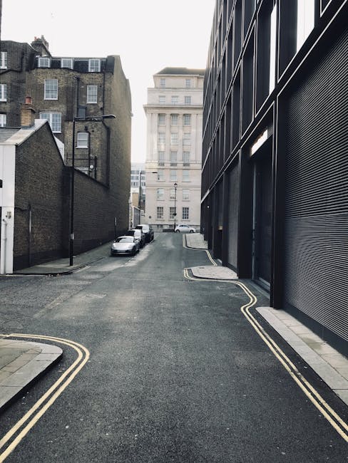A narrow urban street scene showing a steep, one-way road with double yellow lines along the curb edges. On the left side, there are old brick buildings with multiple windows and rooftop structures, while on the right side, a modern black metal-clad building with horizontal slats has an open shutter door revealing part of the interior. Parked cars are lined up along the curb in the distance. The street is mostly clear, with a small stack of cardboard boxes and a large cardboard-wrapped item near the doorway of the modern building, indicating a home relocation process. The overcast sky and muted lighting reflect a typical city environment, suitable for house removals or furniture transport services in Vauxhall, such as those offered by Lambeth Removals, who specialise in careful packing, loading, and moving logistics in tight urban streets.