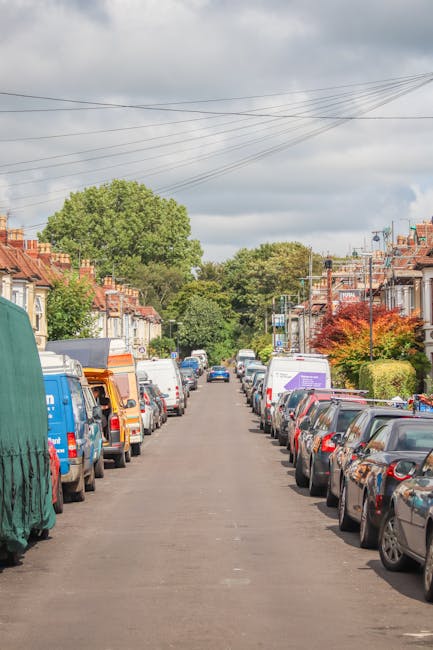 A narrow residential street in Vauxhall lined with parked cars on both sides, including sedans, vans, and small trucks, with some covered in tarps or protective wrapping. The pavement is asphalt, leading toward a cluster of green trees and a blue sky with scattered clouds in the background. Part of a white moving van or lorry belonging to Lambeth Removals is visible on the left side, suggesting the process of home relocation or furniture transport, with the scene capturing a typical street during a loading or unloading process for a house move. Overhead power lines run across the image, and the houses along the street feature brick facades, small front gardens, and pitched roofs, emphasizing the urban residential environment where packing and moving services may be taking place.