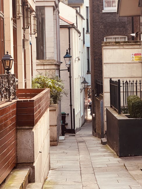 A narrow urban street scene showing a steep, one-way road with double yellow lines along the curb edges. On the left side, there are old brick buildings with multiple windows and rooftop structures, while on the right side, a modern black metal-clad building with horizontal slats has an open shutter door revealing part of the interior. Parked cars are lined up along the curb in the distance. The street is mostly clear, with a small stack of cardboard boxes and a large cardboard-wrapped item near the doorway of the modern building, indicating a home relocation process. The overcast sky and muted lighting reflect a typical city environment, suitable for house removals or furniture transport services in Vauxhall, such as those offered by Lambeth Removals, who specialise in careful packing, loading, and moving logistics in tight urban streets.