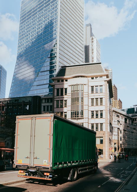 A large green moving truck with closed rear doors is parked on a city street in front of tall modern office buildings with glass facades and older architectural structures. The truck is positioned near the curb, possibly in the process of loading or unloading furniture and packing materials during a home relocation or commercial move. The scene includes a few pedestrians and vehicles on the street, with natural daylight illuminating the area, highlighting the urban environment typical of Lambeth. Lambeth Removals may use similar vehicles for efficient furniture transport and packing and moving services, especially when schedule delays threaten a same-day move, as referenced on the company's webpage.